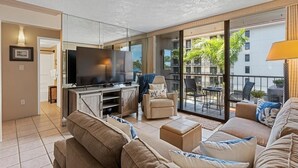 A modern living room with a flat-screen TV, comfortable seating, and a balcony with outdoor furniture. - Pacific Shores A302 3 Bedroom Condo (Kihei)