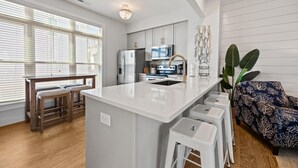 A modern kitchen with white cabinetry, a central island, bar stools, a dining table, and a large plant. - Seas The Day 3 Bedroom Home by RedAwning (Atlantic Beach)