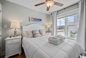 Une chambre à coucher avec un lit, deux tables de chevet avec des lampes, un ventilateur de plafond, une fenêtre avec des rideaux et un tableau au mur.