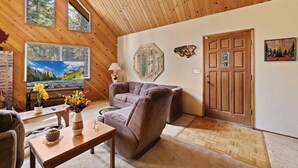A cozy living room with a wooden ceiling, a large window with a mountain view, a sofa, a coffee table with flowers, and a door with a canoe painting.