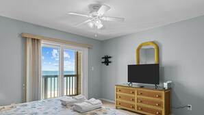 A bedroom with a bed, a television on a dresser, a ceiling fan, and a view of the ocean through a sliding door.