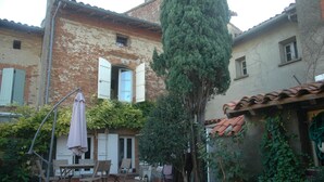 Un patio rústico con un edificio de ladrillo, una mesa y sillas de madera, y un árbol alto.