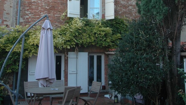 Una casa de ladrillo rústica con un patio que tiene una mesa, sillas, un paraguas grande y un árbol alto.