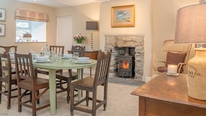 A dining area with a round table set for six, surrounded by wooden chairs. A stone fireplace with a wood-burning stove is visible in the background. - Luxury Farmhouse in Mountain Location | Cwmain (Corwen)