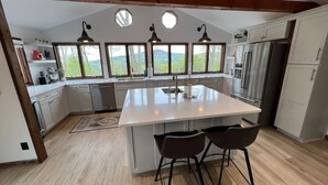 Een moderne keuken met een centraal eiland, houten vloeren en grote ramen met uitzicht op de omgeving.
