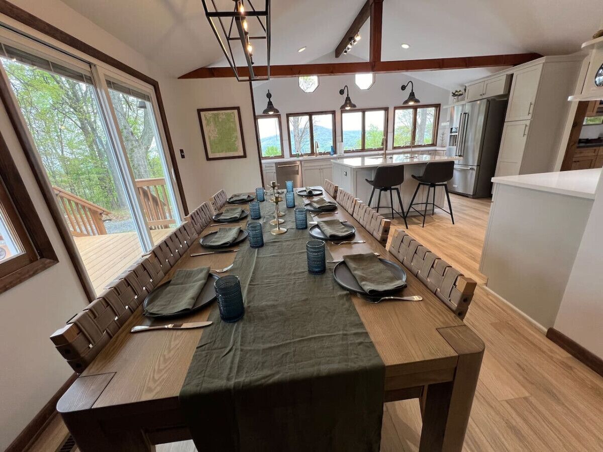 A dining area with a long wooden table set for a meal, a kitchen in the background, and large windows offering a view of the outdoors.