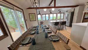 Een eetgedeelte met een lange houten tafel gedekt voor een maaltijd, een keuken op de achtergrond en grote ramen met uitzicht op de omgeving.