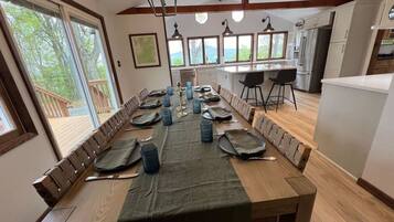 A dining area with a long wooden table set for a meal, a kitchen in the background, and large windows offering a view of the outdoors.