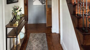Un couloir doté d’un plancher en bois, d’un tapis à motifs, d’un lustre, d’un tableau encadré et d’un escalier orné de balustrades travaillées.