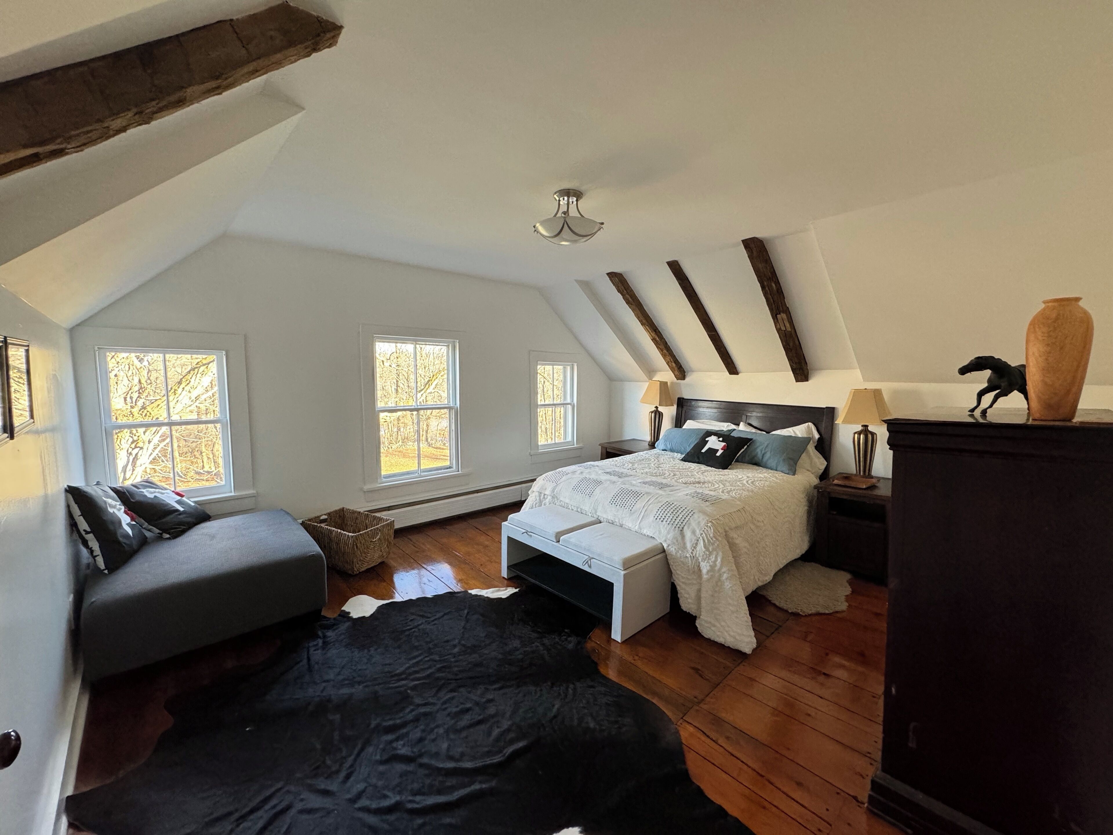Un dormitorio con techo inclinado, una cama con cubrecama blanco, un sofá gris y un tapete de cuero de vaca negro.