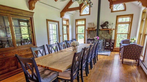 Une grande table à manger en bois, entourée de chaises, avec une cheminée en pierre et de grandes fenêtres en arrière-plan.