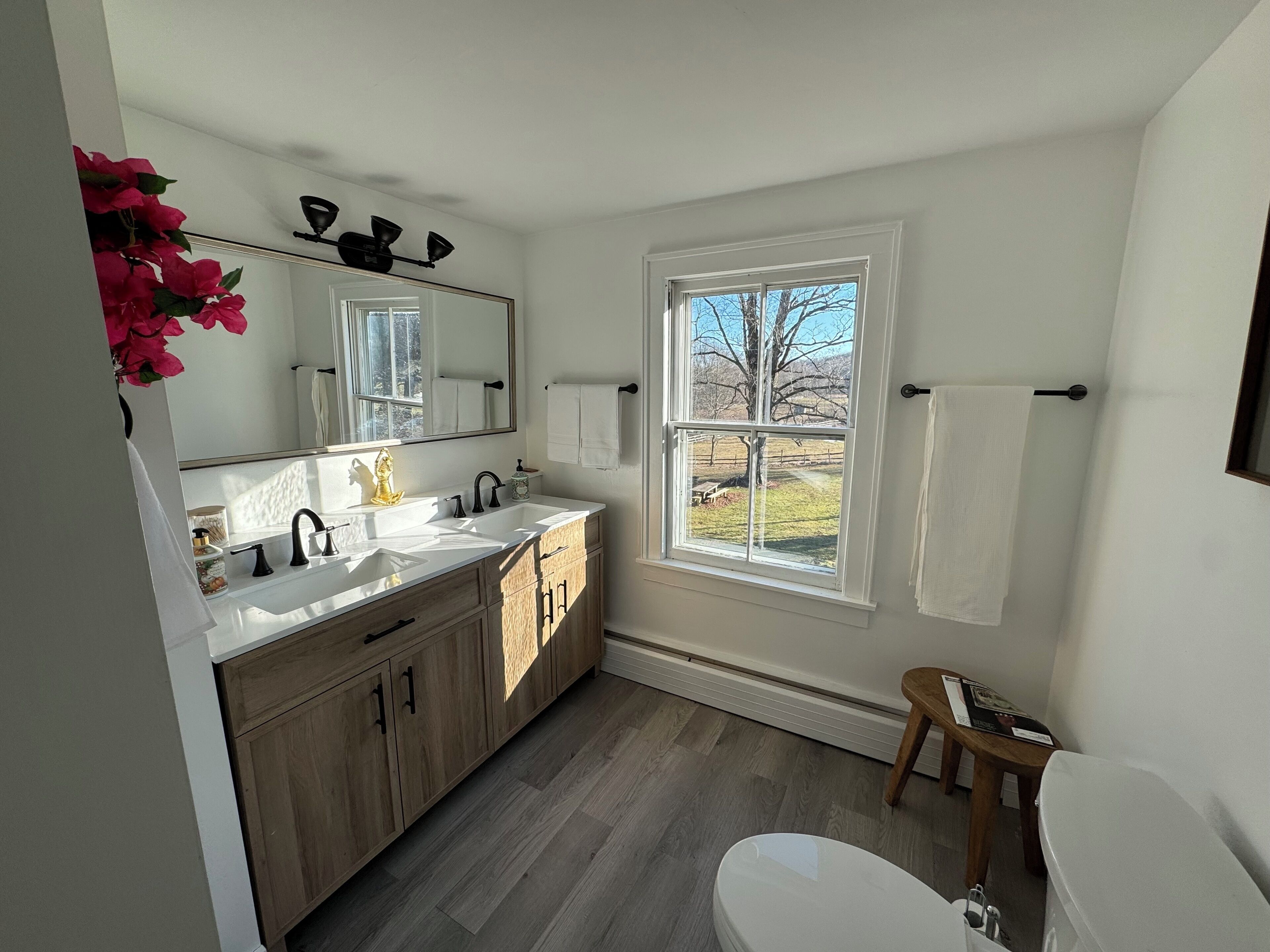 A bathroom with a white sink, wooden cabinets, a large mirror, and a window with a view of a tree and grass.