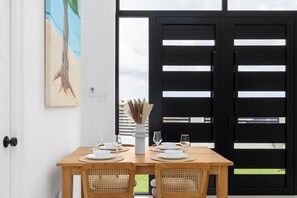 A dining area with a wooden table set for four, surrounded by wicker chairs, a vase with dried plants, and a painting on the wall.
