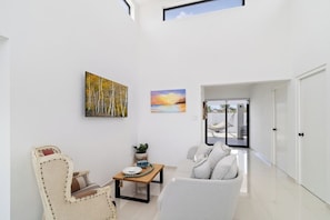 Un salon moderne avec un canapé, un fauteuil et une table basse. Deux tableaux ornent le mur, et une porte donne accès à un espace extérieur.