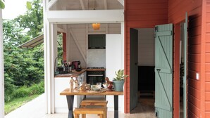 Una mesa de madera con bancos en un espacio exterior cubierto, con vista a una cocina y un edificio rojo.