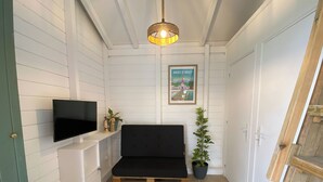 Una habitación pequeña y acogedora con un sofá negro, un televisor montado, un estante de madera con objetos decorativos, un cuadro en la pared y una planta en maceta.