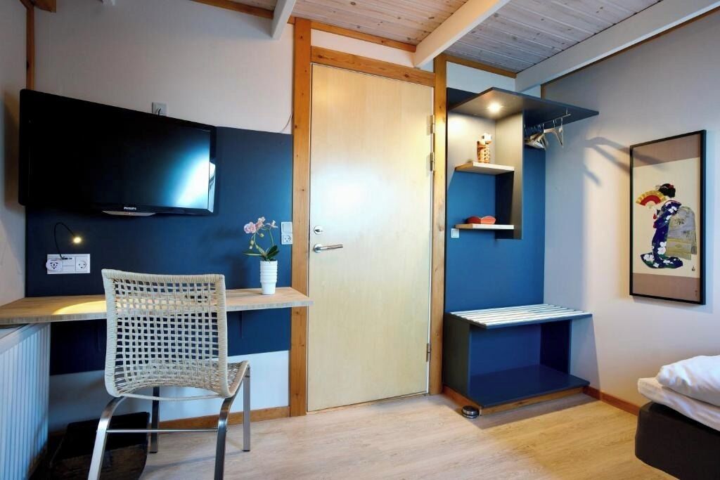 A room with a bed, a desk with a chair, a TV, a door, and a shelf with decorative items.
