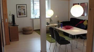 Een moderne woonkamer met een witte tafel, zwarte stoelen, een zwarte bank met rode kussens, een houten kast en een groot raam met jaloezieën.