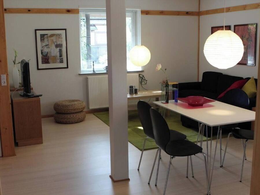 Een moderne woonkamer met een witte tafel, zwarte stoelen, een zwarte bank met rode kussens, een houten kast en een groot raam met jaloezieën.