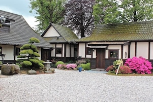 Un edificio de estilo japonés tradicional con techo de tejas, rodeado por un patio de grava, que presenta un bonsái cuidado, un farol de piedra y plantas florecientes y coloridas.