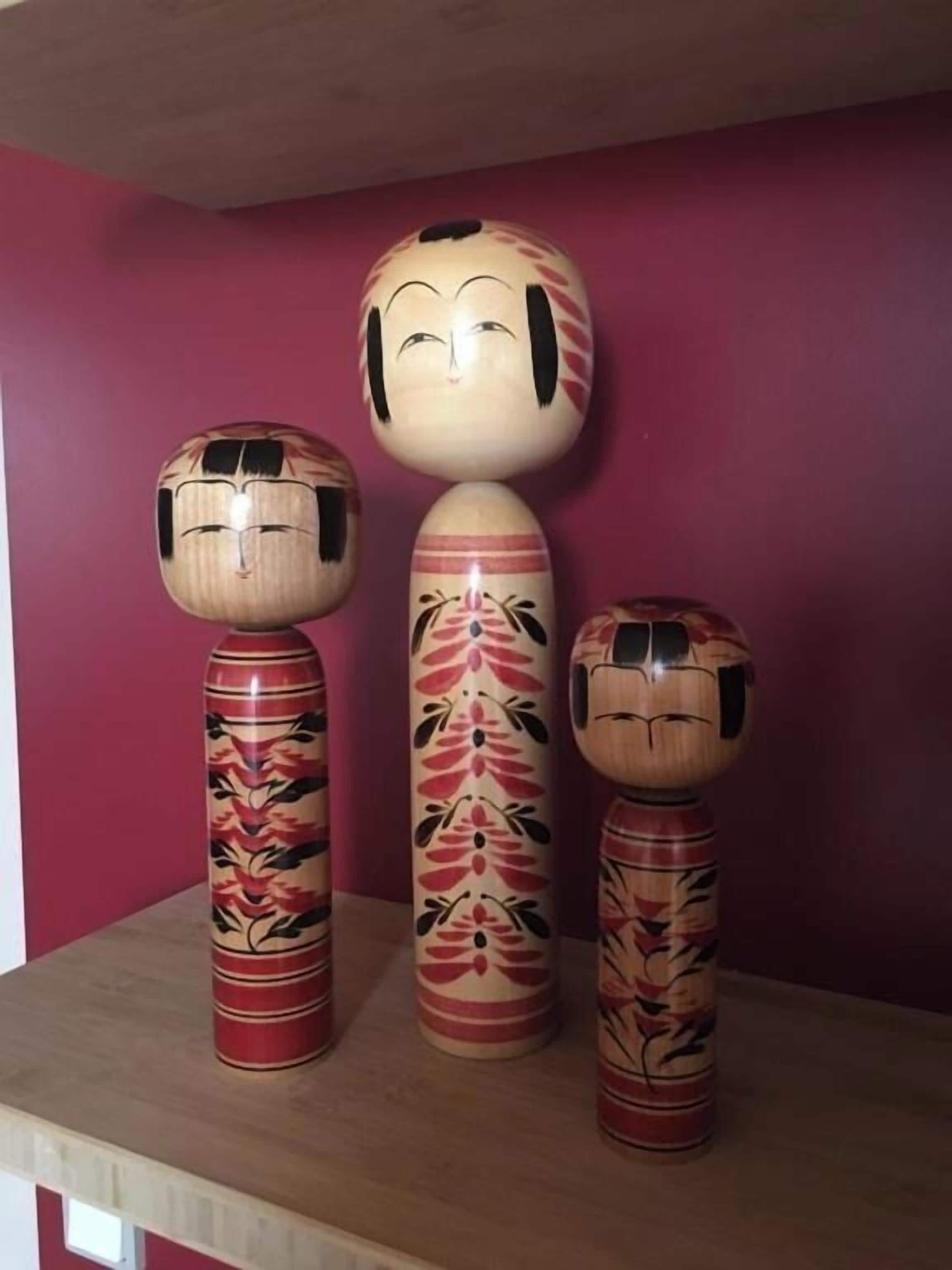 Tres figuras de madera con motivos florales sobre una mesa frente a una pared rosa.