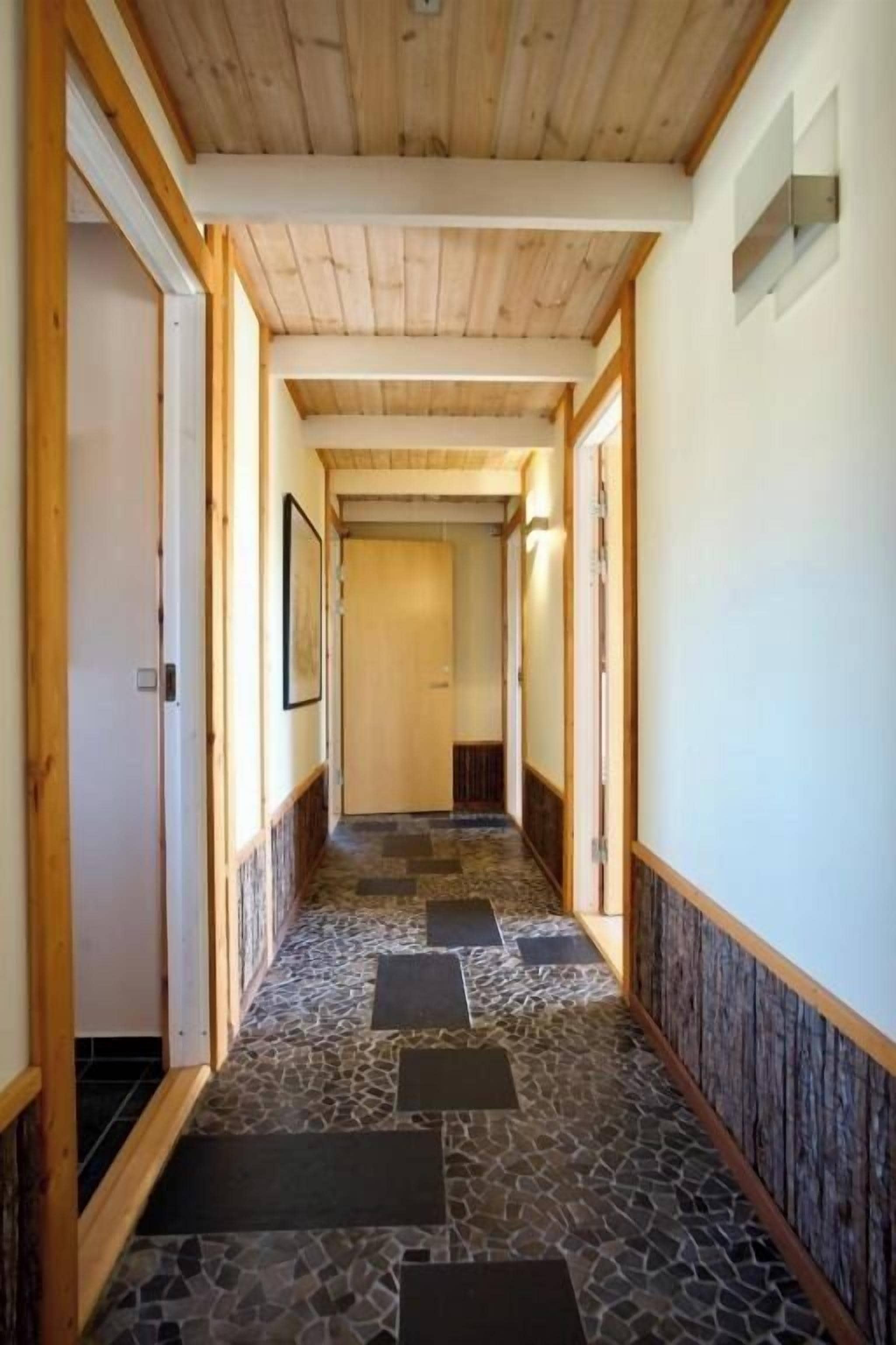 Käytävän katossa on puupalkit ja lattialla kivilaatat, ja se johtaa huoneeseen, jonka päässä on ovi.