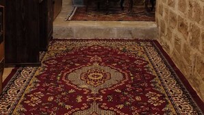 A room with a stone wall, a wooden cabinet, a bed with a headboard, a chair, and a patterned rug. - Rumet Paşa Konağı (Mardin)