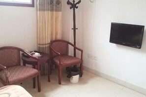 A room with a bed, a TV mounted on the wall, two chairs, a small table, a coat rack, and a window with curtains.