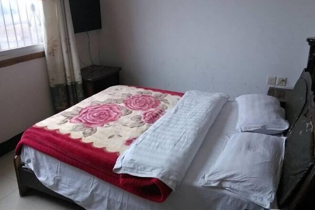 A bed with a floral bedspread, white pillows, and a dark headboard in a room with a window and a wall-mounted TV.