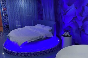 Una cama con colchón y almohadas blancas, una plataforma redonda y una luz ambiental azul.