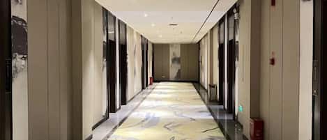 A hotel corridor with a patterned carpet, doors on both sides, and a fire extinguisher on the right.
