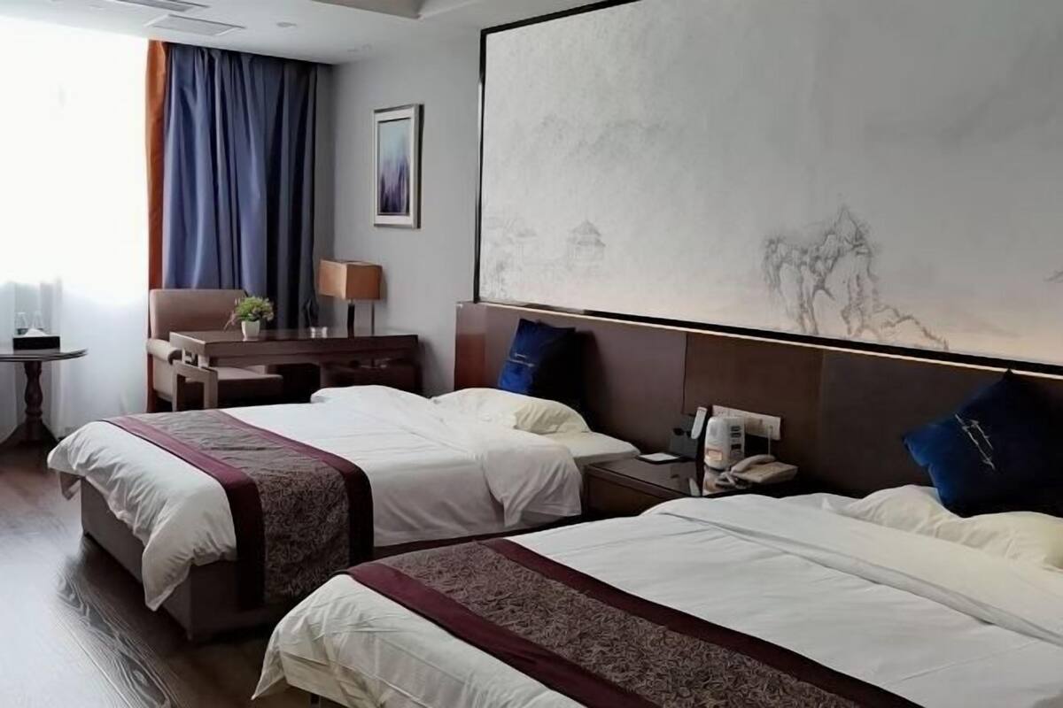 Hotellihuone, jossa on kaksi sänkyä, työpöytä, tuoli, lamppu ja taulu seinällä.