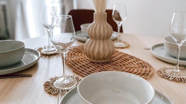Una mesa de comedor dispuesta con platos, tazones y copas de vino, junto con un florero decorativo con plumas.