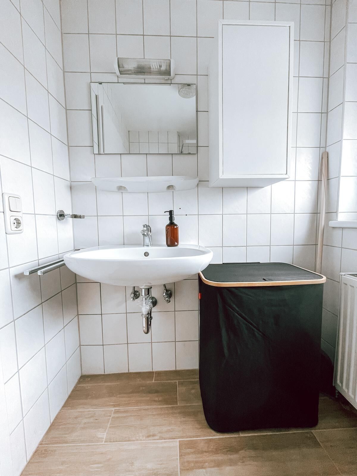 Ein kleines, modernes Badezimmer mit weißen Fliesen, einem weißen Waschbecken, einem Spiegel und einer schwarzen Mülleimer.