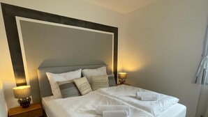 Una cama bien hecha con sábanas blancas y dos toallas enrolladas, una mesita de noche con lámpara y un espejo grande sobre la cama.