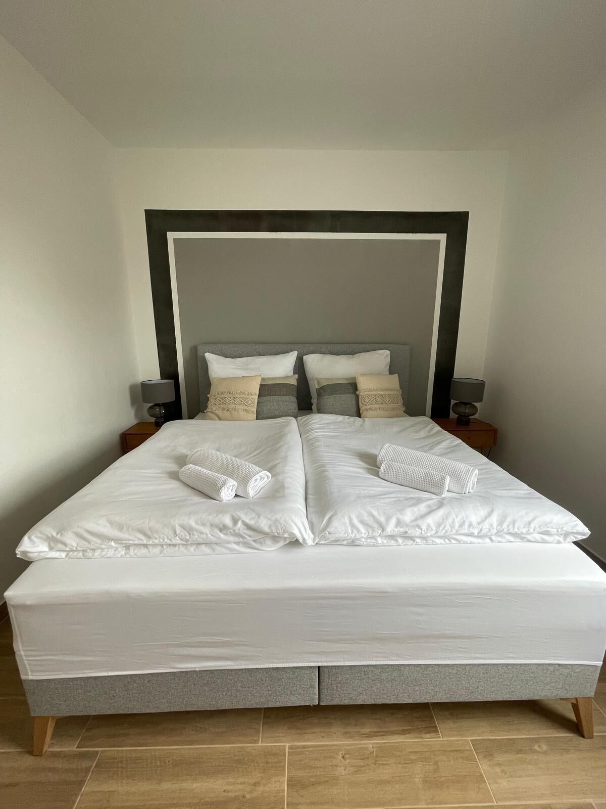 A neatly made bed with white linens and two pillows, a wooden headboard, and two bedside tables with lamps.