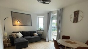 Una sala de estar moderna con un sofá, una mesa de comedor con sillas, un reloj en la pared y un ventanal con cortinas.