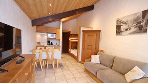 Un ambiente accogliente con un mobile TV in legno, un divano color beige, un tavolo da pranzo con sedie e un angolo cottura con un pensile in legno e un quadro appeso al muro.