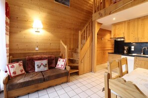 A cozy cabin interior with a wooden staircase, a sofa with decorative pillows, a dining table, and a kitchen area.