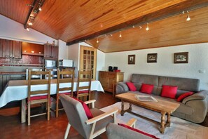 Un salon chaleureux avec un plafond en bois, un canapé gris agrémenté de coussins rouges, une table basse en bois et un coin repas avec des chaises et une table en bois.