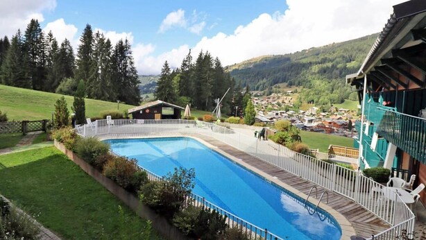 Une piscine dotée d’une terrasse en bois, entourée d’une clôture blanche, avec en toile de fond un paysage montagneux et un petit village.