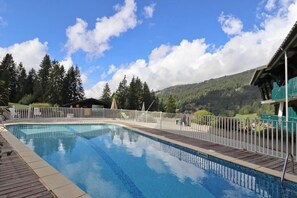 A swimming pool with a wooden deck, surrounded by a white fence, set against a mountainous backdrop.