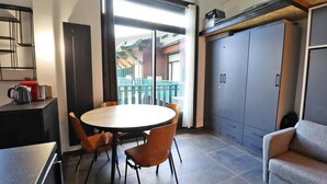 Un espace de vie compact comprenant une table de salle à manger ronde, quatre chaises, un canapé, un coin cuisine et une porte coulissante donnant sur un balcon.