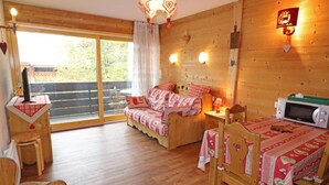 Una habitación acogedora con cama, escritorio y mesa de comedor. Se ve un ventanal con vistas a árboles.