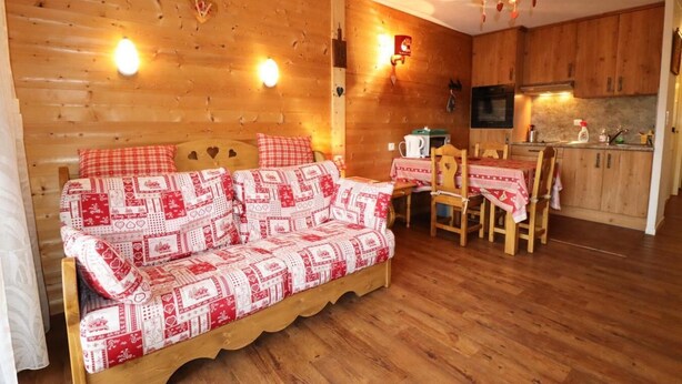 Interior acogedor de una cabaña con sofá de madera, una mesa de comedor con sillas, una zona de cocina y una lámpara colgante.
