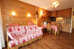 Interior acogedor de una cabaña con sofá de madera, una mesa de comedor con sillas, una zona de cocina y una lámpara colgante.