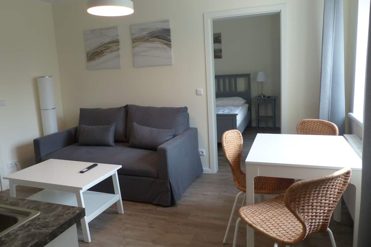 A compact living space with a sofa, a dining table, and two wicker chairs. A bed is visible in the background, and there are two wall-mounted light fixtures.