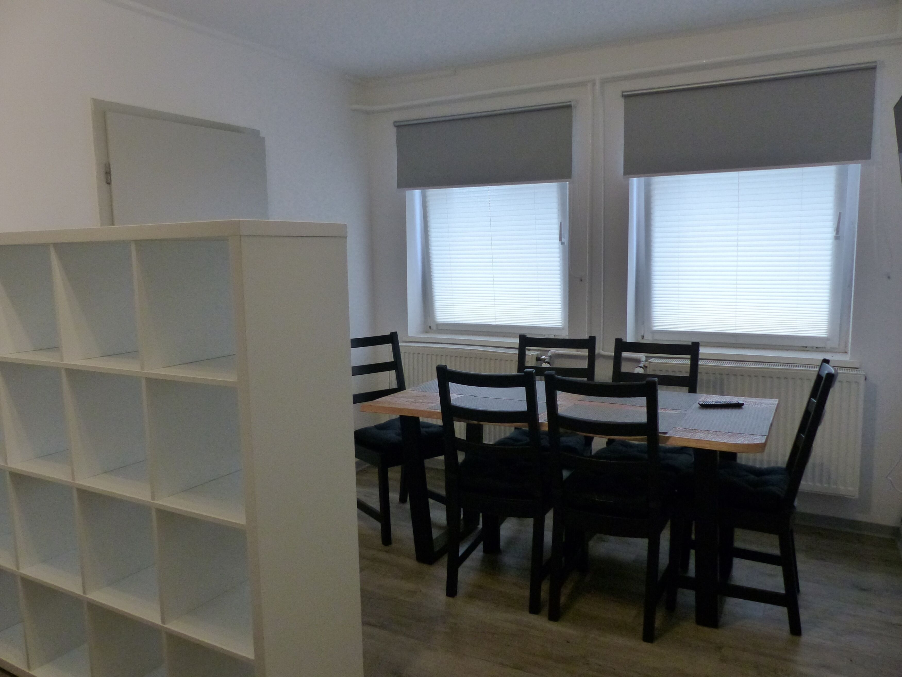 A room with a dining table and chairs, a white shelving unit, and windows with blinds.