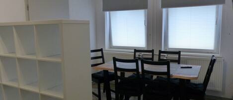 A room with a dining table and chairs, a white shelving unit, and windows with blinds.