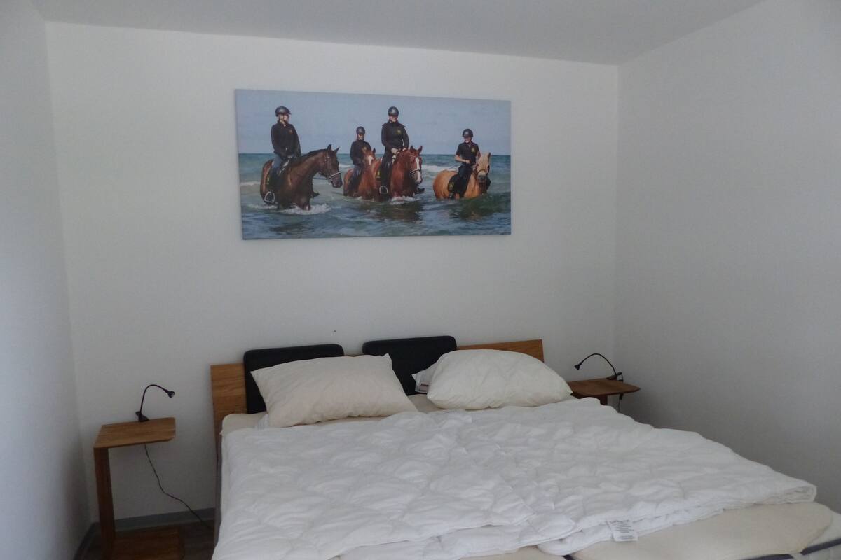 A bedroom with a bed, two bedside tables, a painting of horses on the wall, and a wooden headboard.